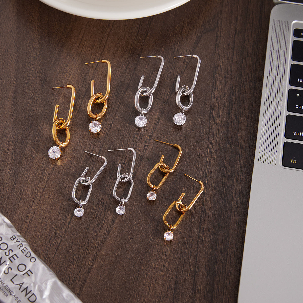 Pair of gold and silver earrings on a wooden surface with a laptop.