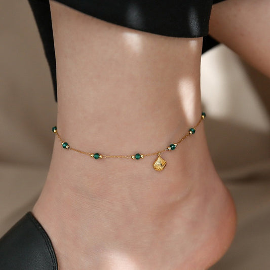 Gold anklet with green beads and a shell charm on a person's ankle against a neutral background