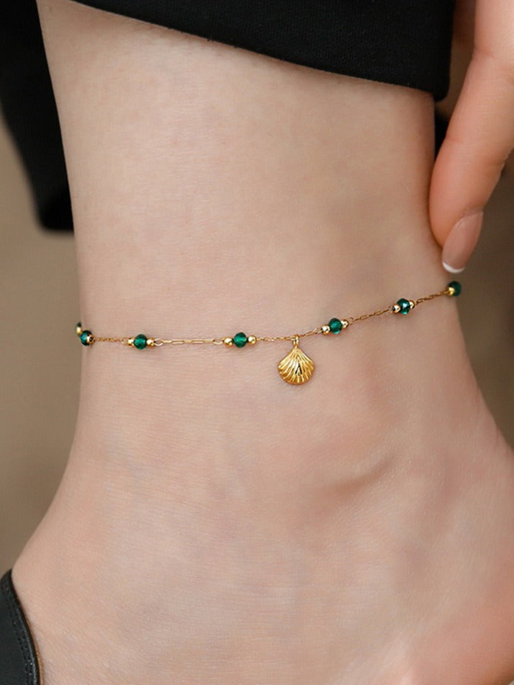 Gold anklet with green beads and a shell charm on a person's ankle.