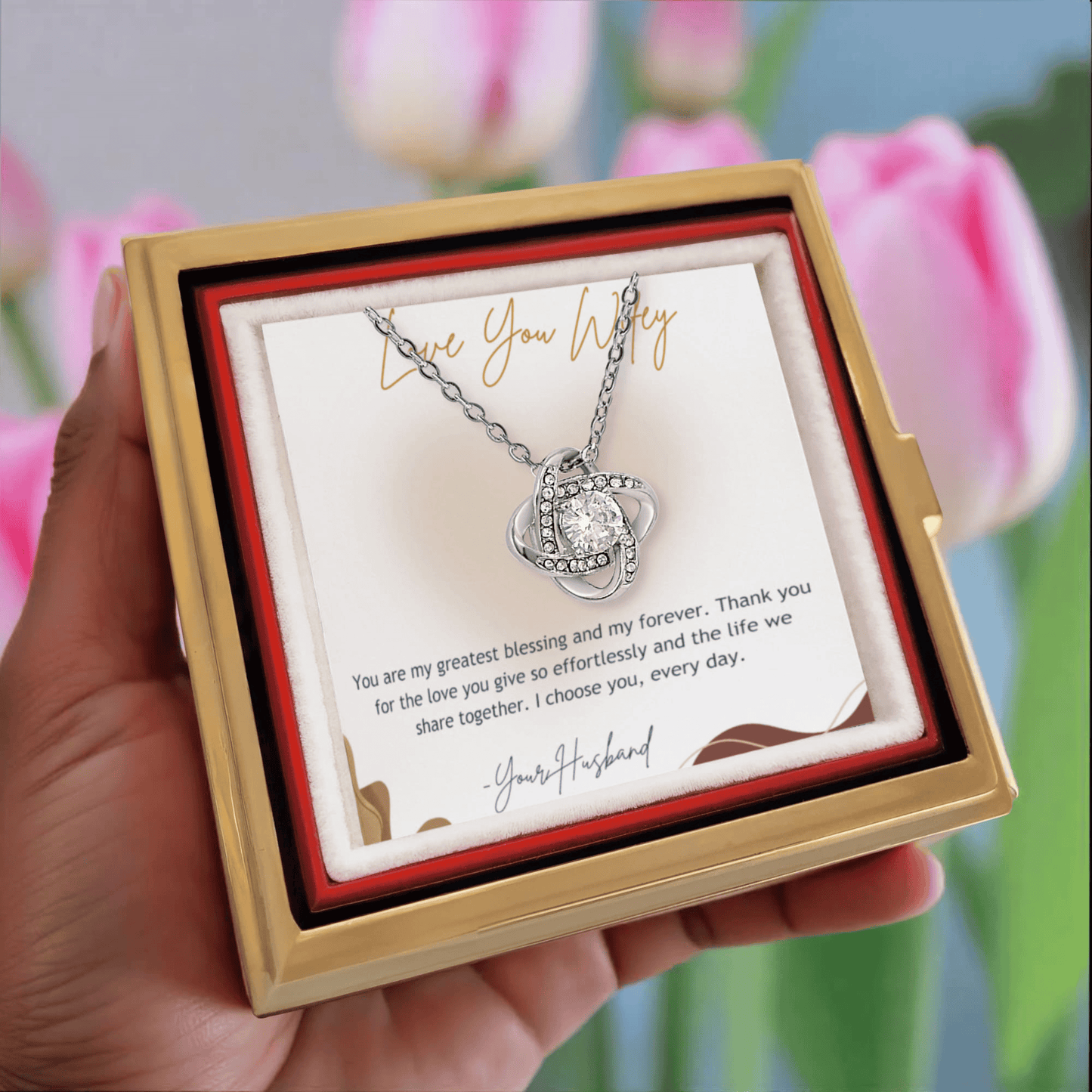 Heart-shaped necklace in a gold box with a card, held against a blurred tulip background.