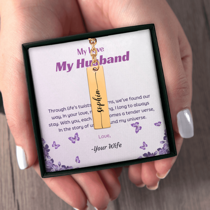 Gold necklace with a tag in a box labeled 'My Love, My Husband' held by a hand.