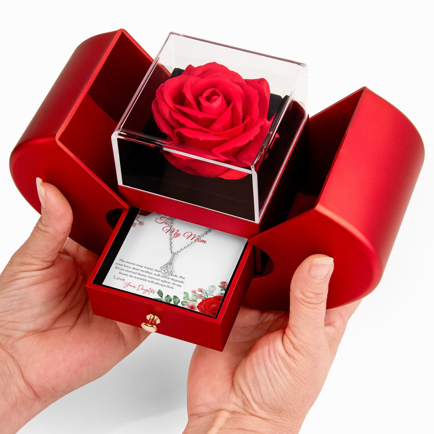 Red rose in a clear cube within a heart shaped red box with a necklace and card held by a hand on a white background